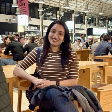 Ipshita is an Indian woman with long, dark hair with subtle burgundy highlights and a warm smile. She is wearing a brown-and-white striped top and is seated at a wooden table inside the lively Time Out Market in Lisbon, Portugal, with restaurants and people dining in the background.