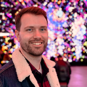 David smiles brightly in front of a glittering background of multi-hued purple lights. He’s now in his thirties, a Scottish-Canadian with short brown hair.