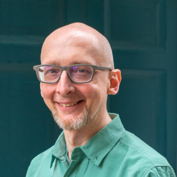Gary Morgan is a white man with a shaved head and dark green glasses, wearing a bright green shirt.