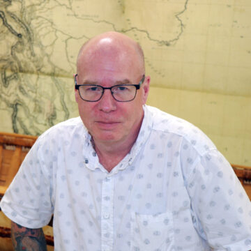 Hair challenged caucasian male wearing glasses and a short-sleeved white button-up shirt, sitting in front of a old map.