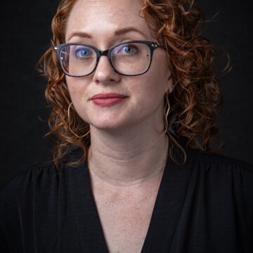 Katie is a curly redhead, white woman, wearing a black shirt, glasses, and a slight smile