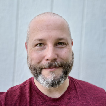 Chris Hinds is a white man in his early 40's with a bald head and a salt-and-pepper trimmed beard. He has a slight smile and friendly eyes.