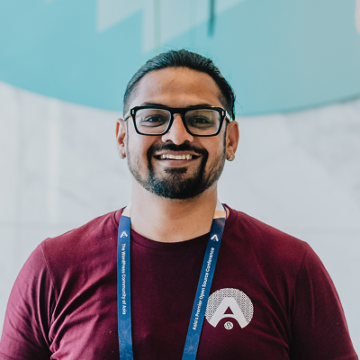 Mayank Majeji is an Indian man with medium-brown skin, dark hair pulled back, black glasses, and a short beard. He is smiling at the camera, wearing a maroon T-shirt and a blue WordCamp Asia 2026 lanyard.