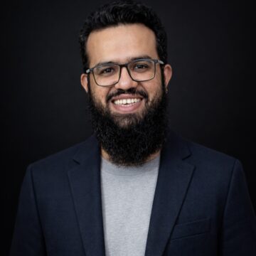 Muhammad Asad is a Pakistan man, smiling in a professional portrait, wearing glasses, a dark blazer, and gray shirt against a dark background.