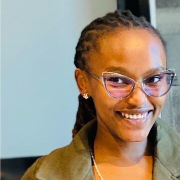 Rose is a black woman in her 20's with glasses and dreadlocks smiling at the camera