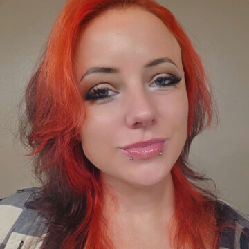 Kadian is a feminine-looking caucasian person in their mid-thirties. They have a fire-engine red halo with a brunette crown of mid-length wavy hair, a dark, smokey eye makeup, and two silver lip piercings ("snakebites"). They are wearing a black-and-gray plaid shirt and smiling slightly at the camera.