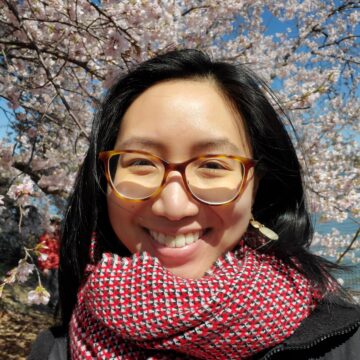 Angela Fong is an East Asian woman with black hair, turotise shell glasses, smiling in front of cherry blossoms in bloom.
