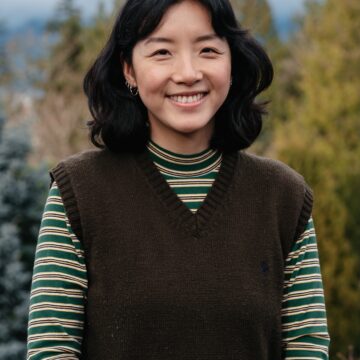 Hailey Mah is a Chinese woman with wavy shoulder-length black hair and silver hoop earrings. She is wearing a green sweater vest and green striped turtleneck, and stands outside with trees and mountains out of focus in the background.