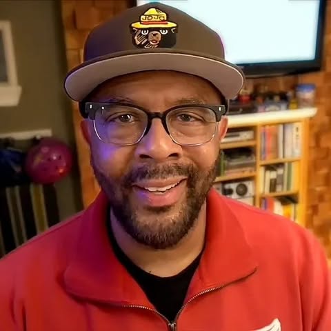 Joe Simpson Jr. is smiling, wearing black-framed glasses, a dark brown baseball cap, and a red zip-up jacket.